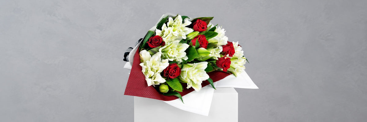 A bouquet of red roses, white rose lilies, and white carnations wrapped in a dark red and white paper sleeve, displayed on a white pedestal against a textured gray background.